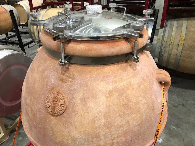 Large traditional clay fermentation vessel used in winemaking, with metal fittings and a glass lid, in a winery setting.
