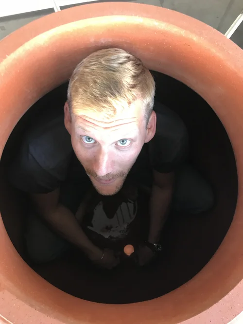 A man with blond hair and blue eyes looking up from inside a large terracotta pot.