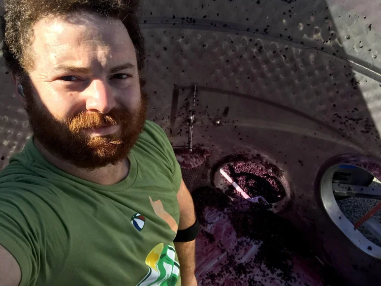 A man with a beard taking a selfie inside a wine barrel, wearing a green shirt with a logo, surrounded by wine-making equipme.