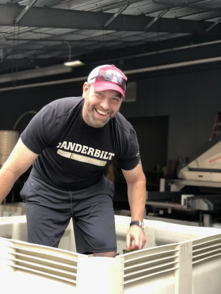 Happy man inspecting wine barrels in a winery, wearing a black t-shirt and red cap, smiling at the camera.