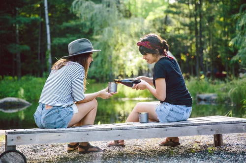 A woman in a striped shirt and hat offers a cup to a woman in a black shirt and headband, sitting on a wooden bench by a lake.